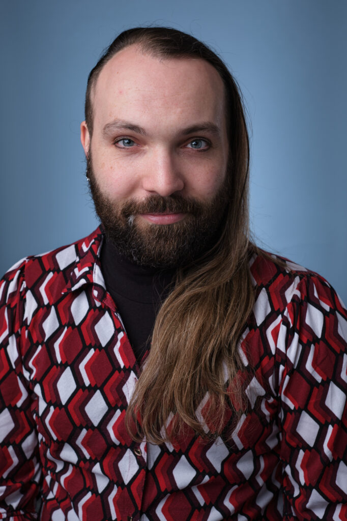 Photo of Jordan Rosin, a white, nonbinary person wearing a bright red shirt, with long brown hair and short brown beard.