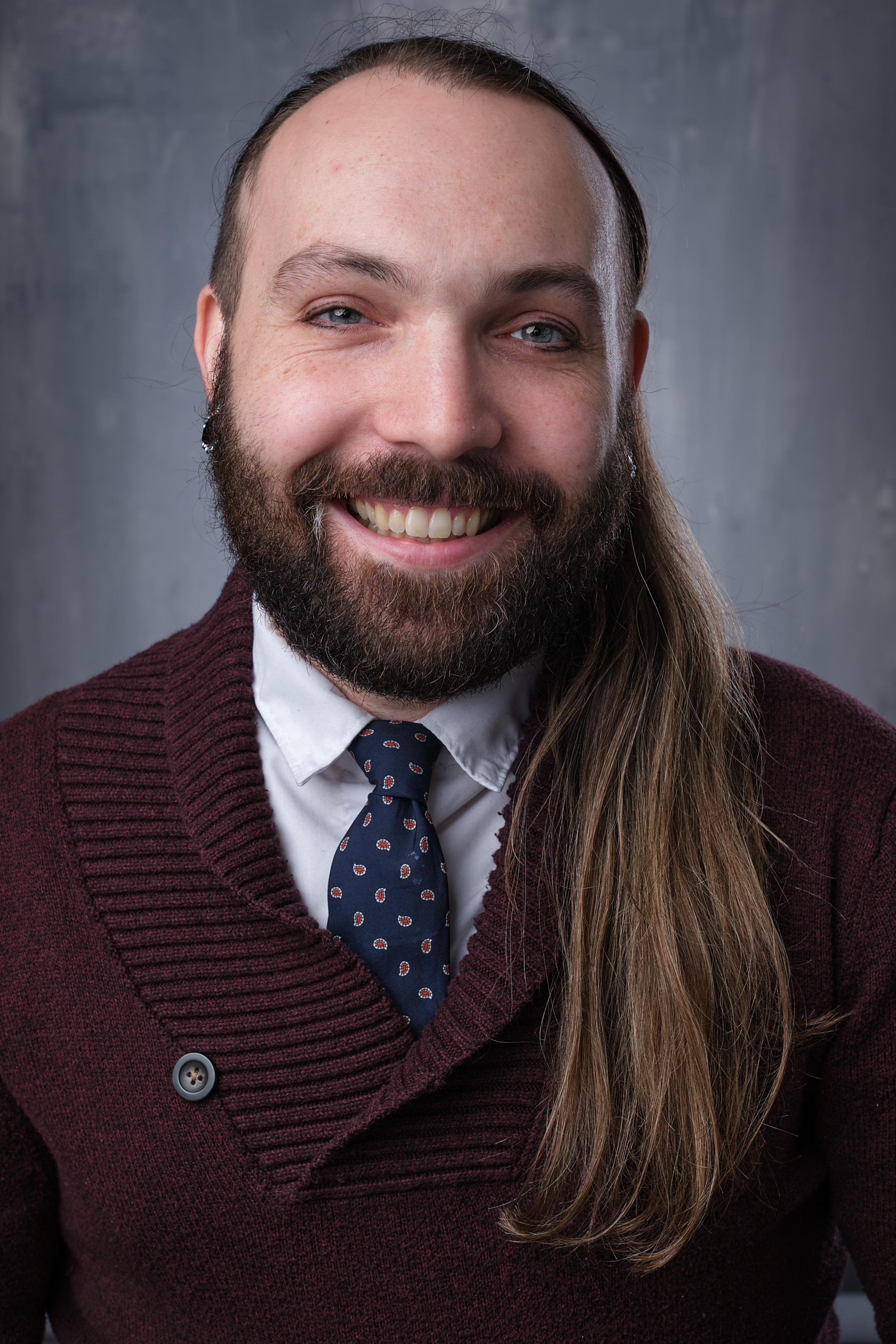 Photo of Jordan Rosin, a white, nonbinary person wearing a a white collared shirt with blue tie under a maroon sweater, with long brown hair and short brown beard.
